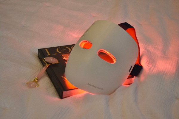 TheraFace Mask Glo On Bed red light mask on a book on a bed white a gua sha on light gray background